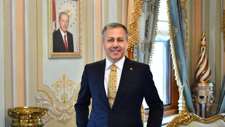 Takım elbiseli bir kişi, süslü bir salonda; arka planda duvarda portre ve bayraklar görülüyor.