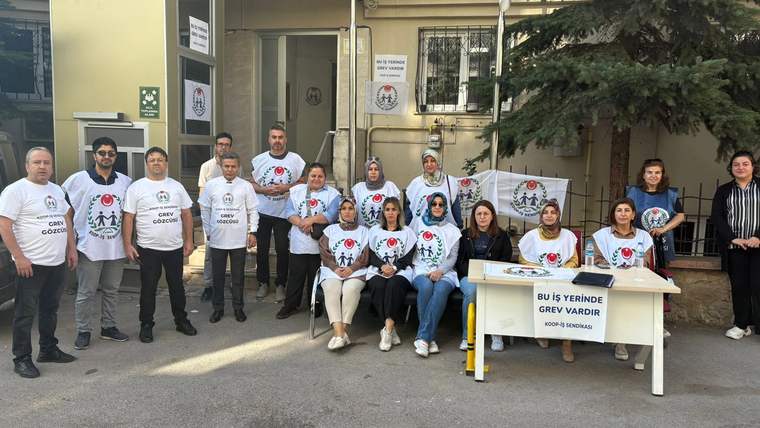 Bir bina önünde, üzerinde yazılar bulunan beyaz tişörtlü kişiler ve grev yazılı bir masa görülüyor.