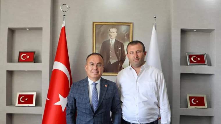 Türk bayrakları ve Atatürk portresi önünde duran iki erkek, duvar raflarındaki plaketlerle birlikte görülüyor.