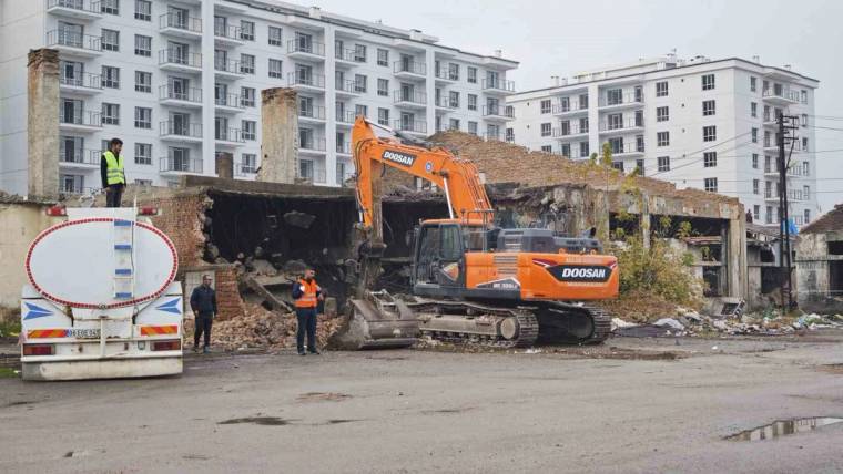 Turuncu ekskavatör, yıkılmış bir yapının enkazı önünde çalışırken çevrede işçiler ve su tankeri görülüyor.