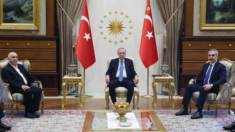 Ortada koltukta oturan bir kişi, iki yanında sandalyelerde oturan kişiler ve arkalarında Türk bayrakları bulunuyor.