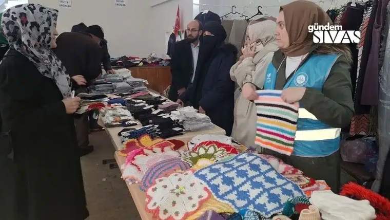 Kapalı salonda el işi ürünlerle dolu masanın çevresinde duran kişiler ve kıyafet askıları görülüyor.