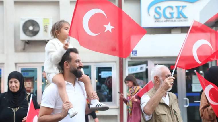 Sokakta Türk bayrakları taşıyan kişiler arasında, omzunda çocuk taşıyan bir adam ve yaşlı bir adam görülüyor.