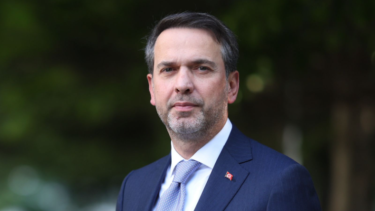 Koyu mavi takım elbise giymiş bir adam, dış mekanda duruyor ve kameraya bakıyor.