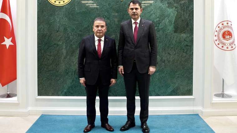 İki kişi, yeşil arka planda yan yana duruyor. Biri siyah takım elbise, diğeri ise koyu mavi takım elbise giymiş.