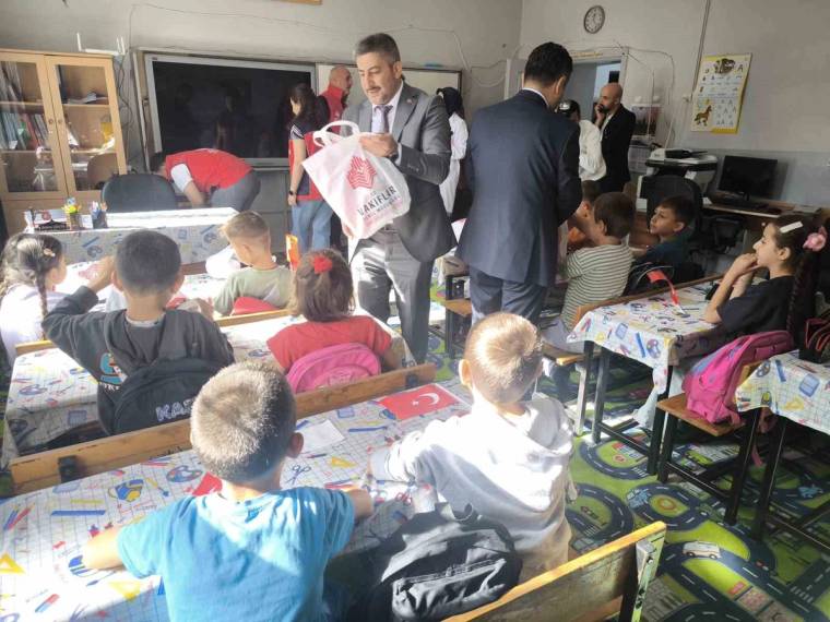Bir sınıfta masalarda oturan çocuklar, ayakta duran yetişkinler ve ellerinde poşet bulunan bir kişi görülüyor.