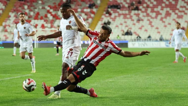 Kırmızı-beyaz formali futbolcu, rakibiyle mücadele ederken çim sahada topa uzanıyor; diğer oyuncular izliyor.