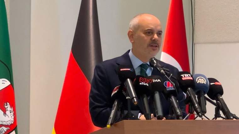 Kürsüde konuşan bir kişi, önünde çok sayıda mikrofon ve arkasında Türk bayrağıyla duruyor.