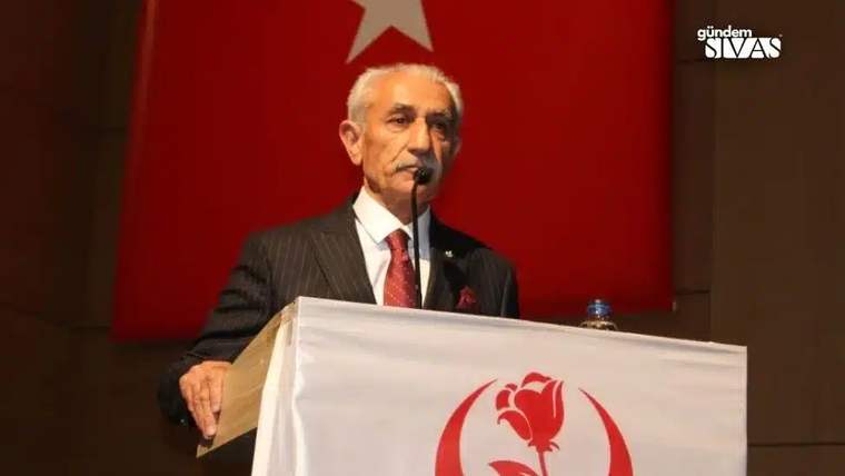 Kürsüde konuşan bir kişi, arkasında Türk bayrağı ile birlikte bir pankart var.