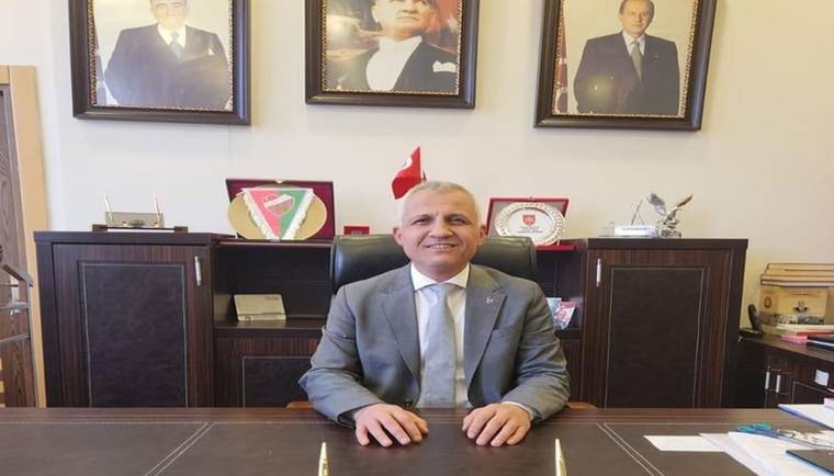 Takım elbiseli bir kişi, arkasındaki portreler ve Türk bayrağı önünde makam masasının başında oturuyor.