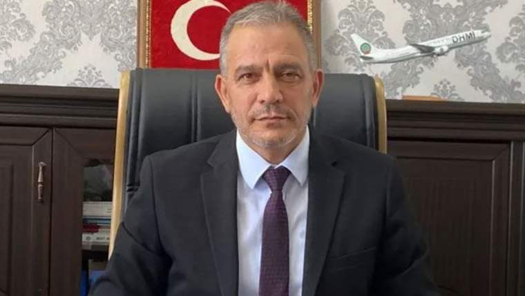 Koltukta oturan bir kişi, takım elbise giymekte ve arkasında Türk bayrağı ile DHMİ logosu görünmekte.
