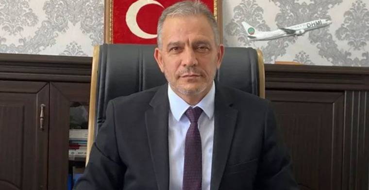 Koltukta oturan bir kişi, takım elbise giymekte ve arkasında Türk bayrağı ile DHMİ logosu görünmekte.
