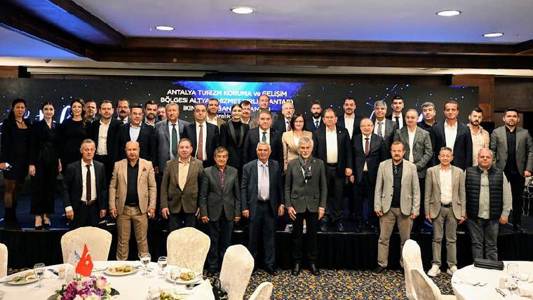Bir otelde düzenlenen etkinlikte, katılımcılar grup halinde poz veriyor. Arka planda tanıtım panosu var.