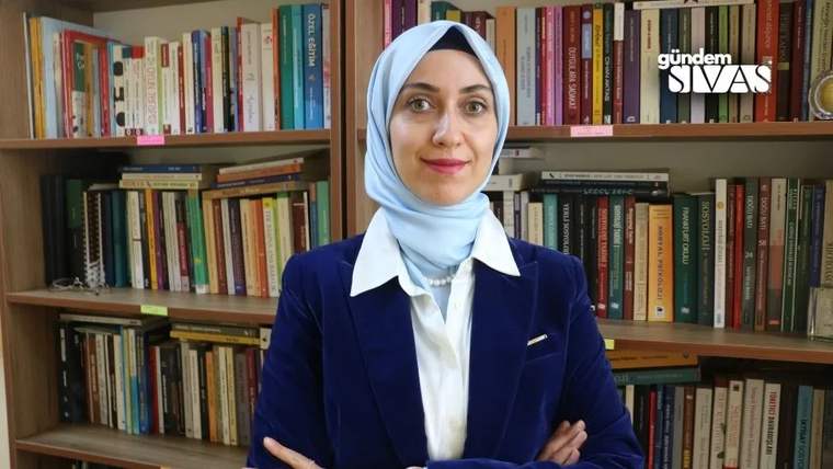 Başörtülü bir kadın, lacivert ceket ve beyaz gömlekle kitap raflarının önünde kollarını bağlamış duruyor.
