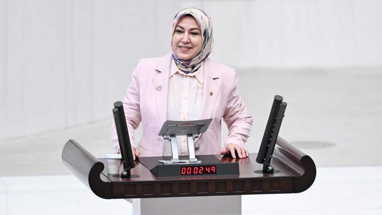 Konuşma yapan bir kadın, kürsüde duruyor ve arkasında beyaz bir duvar var.