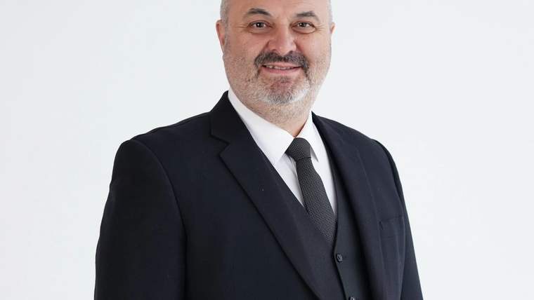 Siyah takım elbise giymiş, gülümseyen bir adam, beyaz arka planda duruyor.