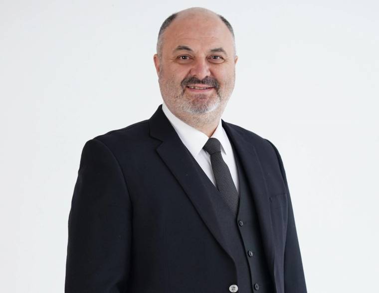 Beyaz arka plan önünde koyu renk takım elbiseli, sakallı ve gülümseyen bir erkek portresi.