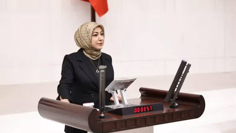 Kürsüde konuşan başörtülü bir kadın, önündeki ekranda 000351 yazan salonda görülüyor.