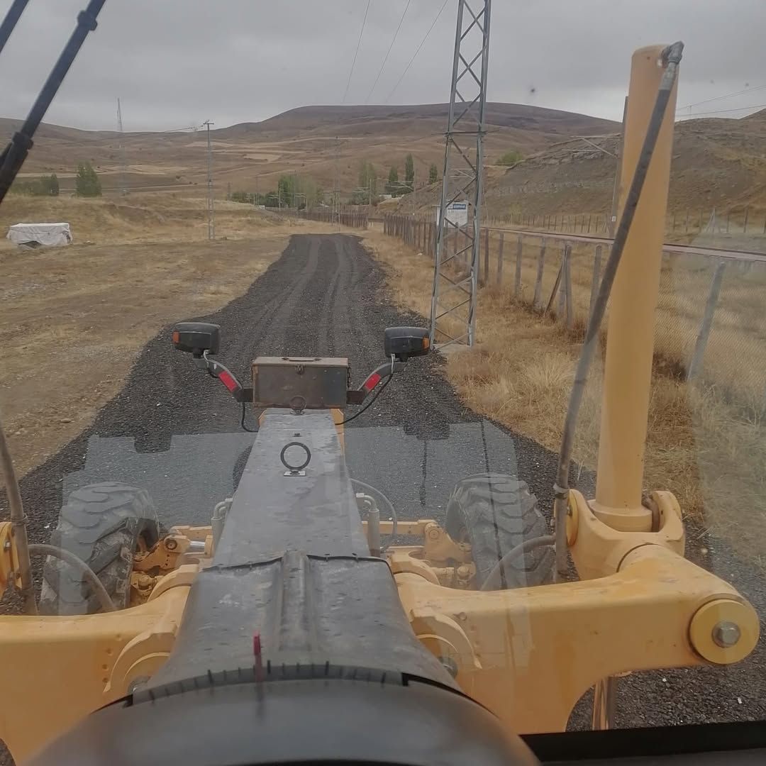 Sarı bir iş makinesi, kırsal alanda siyah malzemeyle kaplanan yolun üzerinde görülüyor.