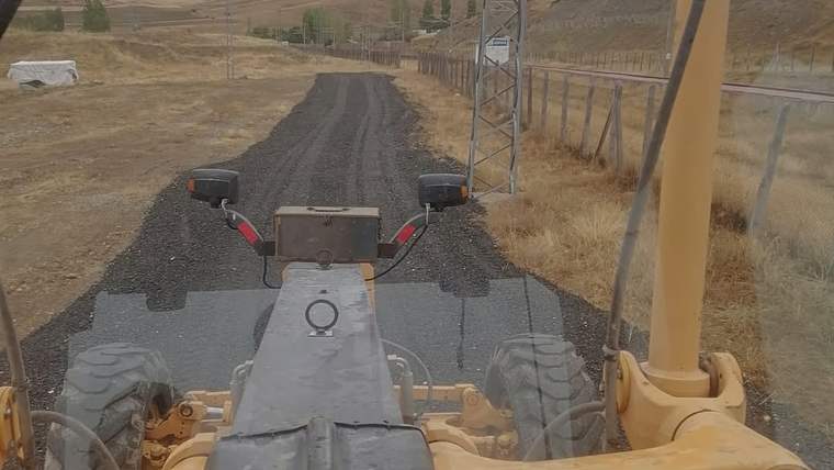 Sarı bir iş makinesi, kırsal alanda siyah malzemeyle kaplanan yolun üzerinde görülüyor.