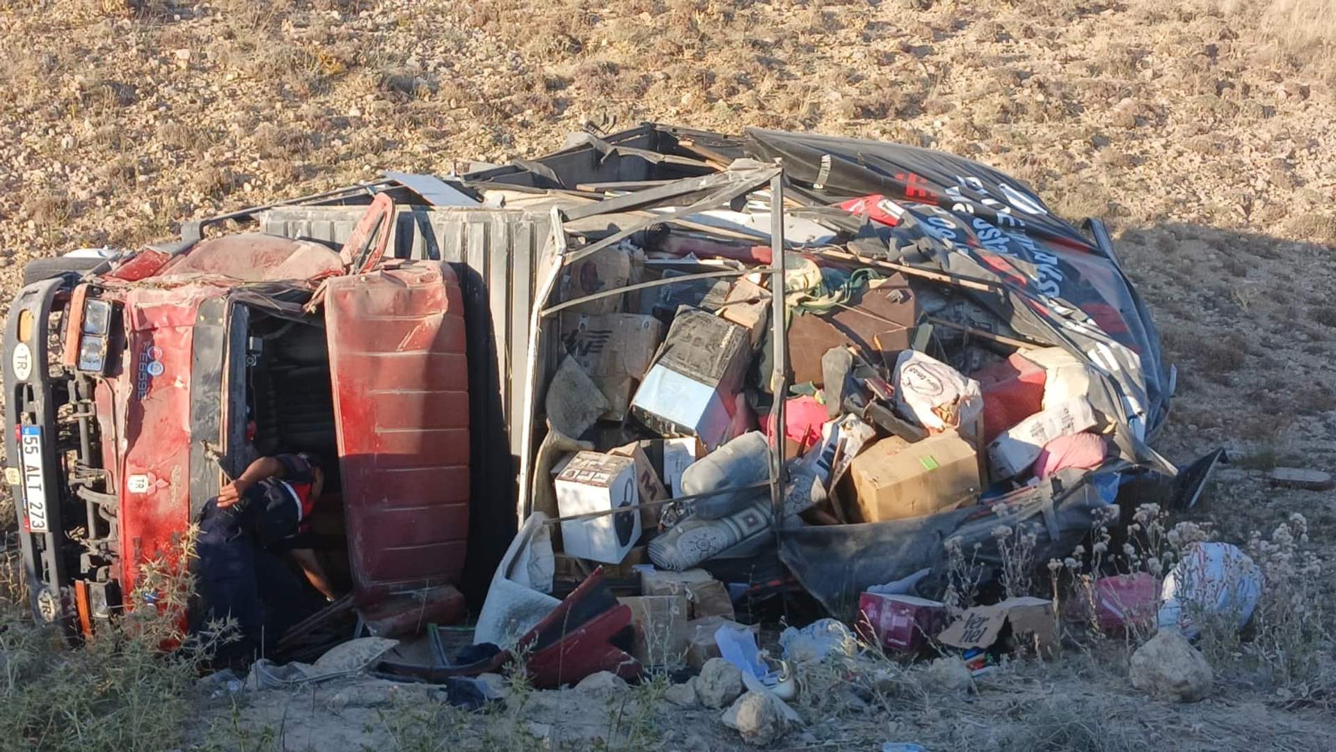Yol kenarındaki kuru arazide yan yatmış, içindeki eşyalar çevreye saçılmış kırmızı bir araç görülüyor.