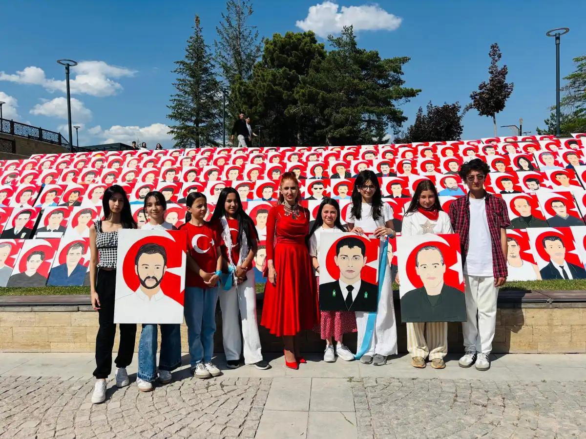 Açık havada bir grup genç, ellerinde portre çizimleriyle kırmızı-beyaz portrelerle kaplı pano önünde duruyor.