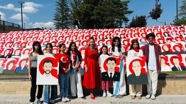 Açık havada bir grup genç, ellerinde portre çizimleriyle kırmızı-beyaz portrelerle kaplı pano önünde duruyor.