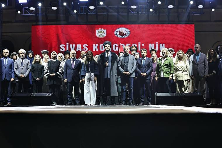 Sahnede çok sayıda kişi yan yana duruyor; arkadaki kırmızı ekranda “Sivas Kongresi” yazısı görülüyor.