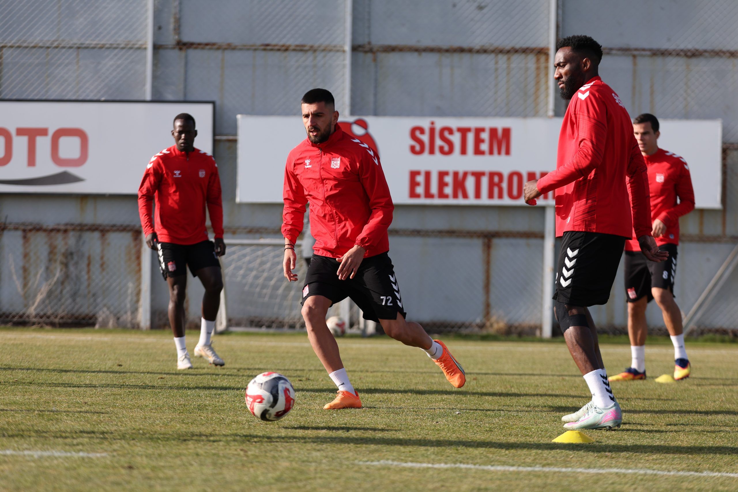 Çim sahada kırmızı formalar giyen beş futbolcu, antrenman sırasında bir topun çevresinde koşuyor.
