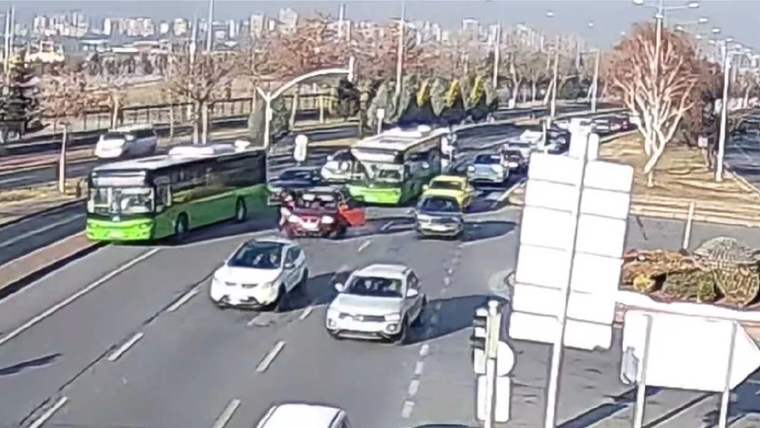 Çok şeritli kavşakta yeşil belediye otobüsleri, otomobiller, trafik ışıkları ve ağaçlar görülüyor.