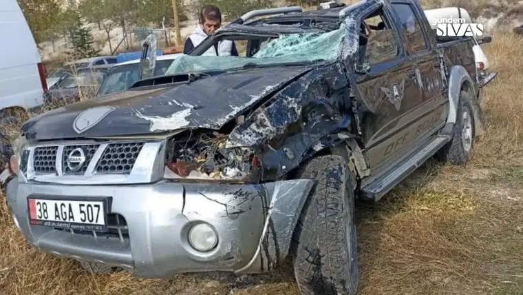 Ön kısmı ağır hasar gören Nissan araç, kırılmış camlarıyla çimenlik alanda duruyor; yanında bir kişi var.