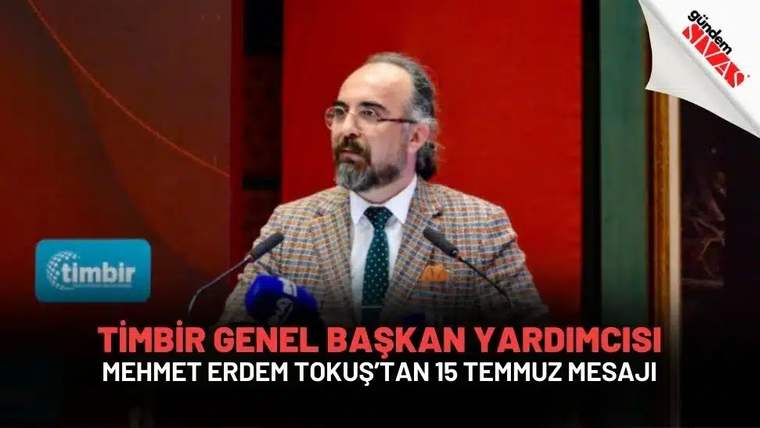 Gözlüklü, sakallı bir kişi iki mikrofon önünde konuşurken; arka planda kırmızı panel ve kurum logosu görülüyor.