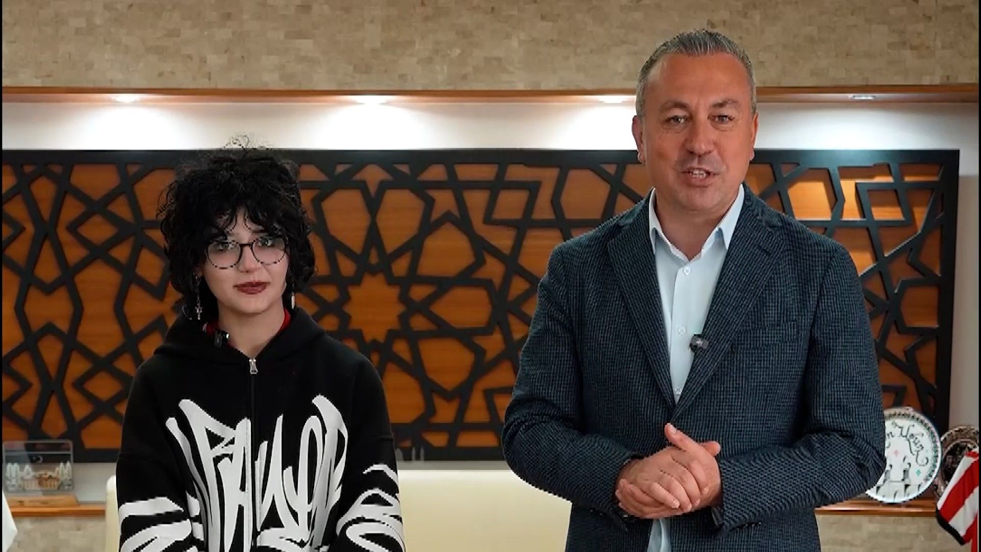 Bir kişi, yanında başka bir kişi ile birlikte gülümseyerek konuşuyor. Arka planda dekoratif bir duvar var.