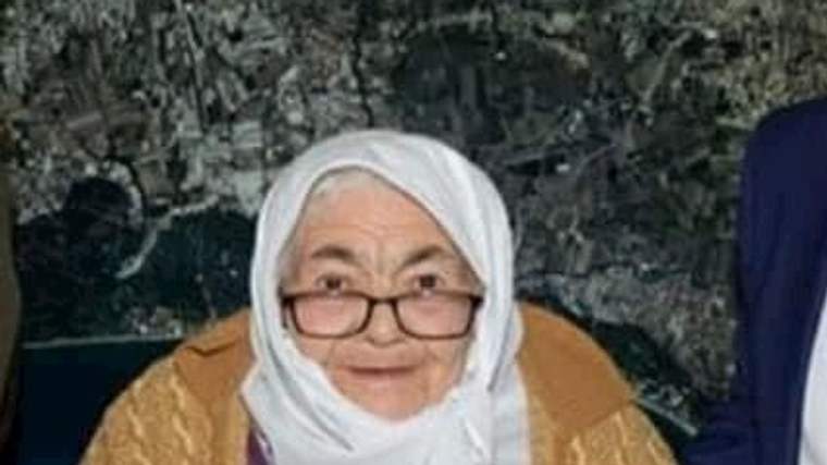 Beyaz başörtülü, gözlüklü yaşlı kadın mor kıyafet ve kahverengi hırkayla otururken görülüyor.