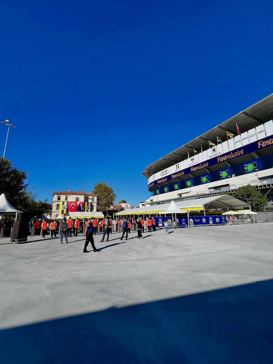 Fenerbahçe yazılı stadyumun önünde Türk bayrakları ve çeşitli bayraklar arasında insanlar bekliyor.