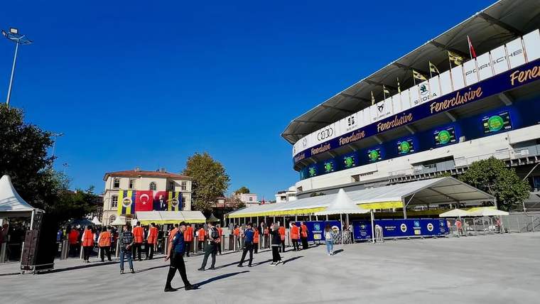 Fenerbahçe yazılı stadyumun önünde Türk bayrakları ve çeşitli bayraklar arasında insanlar bekliyor.