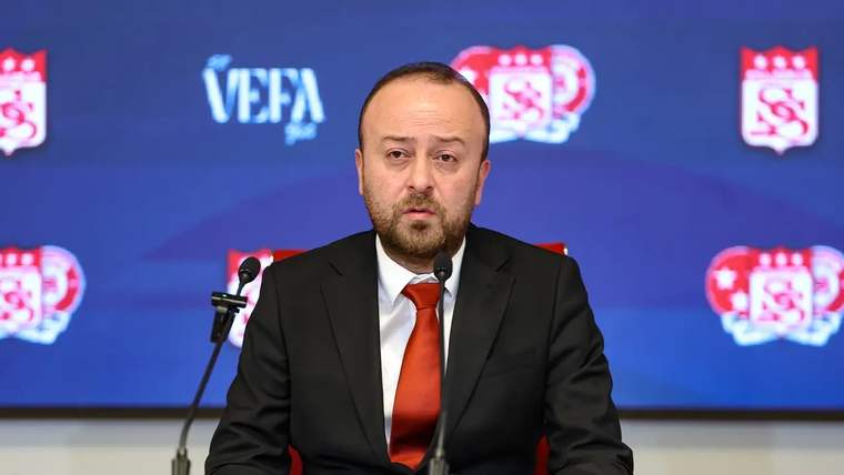 Kırmızı kravat takan bir kişi, mikrofonun önünde oturarak açıklama yapıyor.