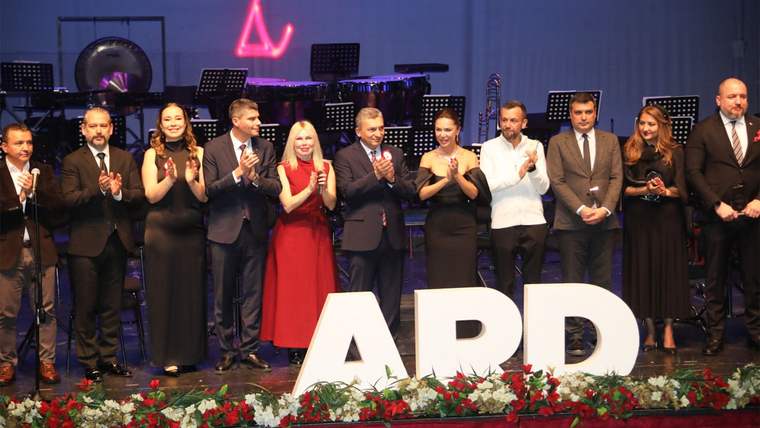 Sahnede yan yana duran dokuz kişi alkışlıyor; önlerinde büyük beyaz harflerle “ARD” yazısı ve çiçekler bulunuyor.