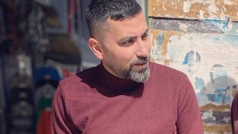 Kısa saçlı ve sakallı bir erkek, bordo kazakla dışarıda bir yere bakarken görülüyor.