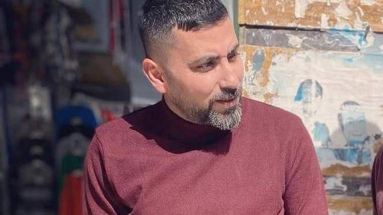 Kısa saçlı ve sakallı bir erkek, bordo kazakla dışarıda bir yere bakarken görülüyor.