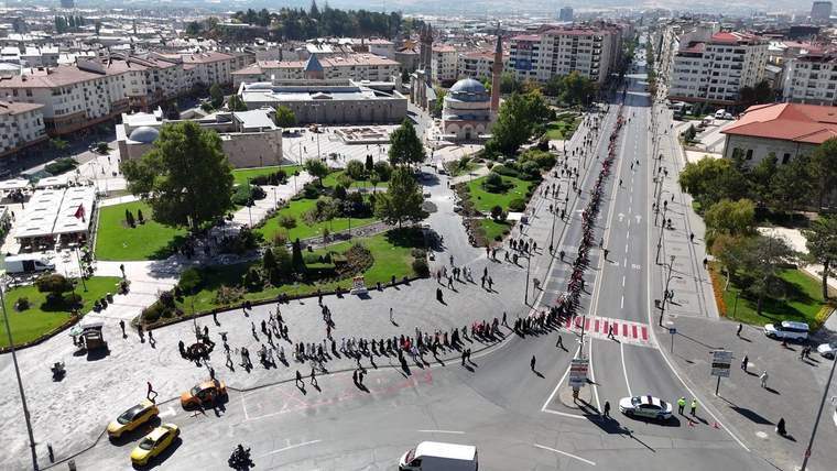 Şehir merkezinde cami, park, çok katlı binalar ve caddede sıralar halinde ilerleyen kalabalık görülüyor.