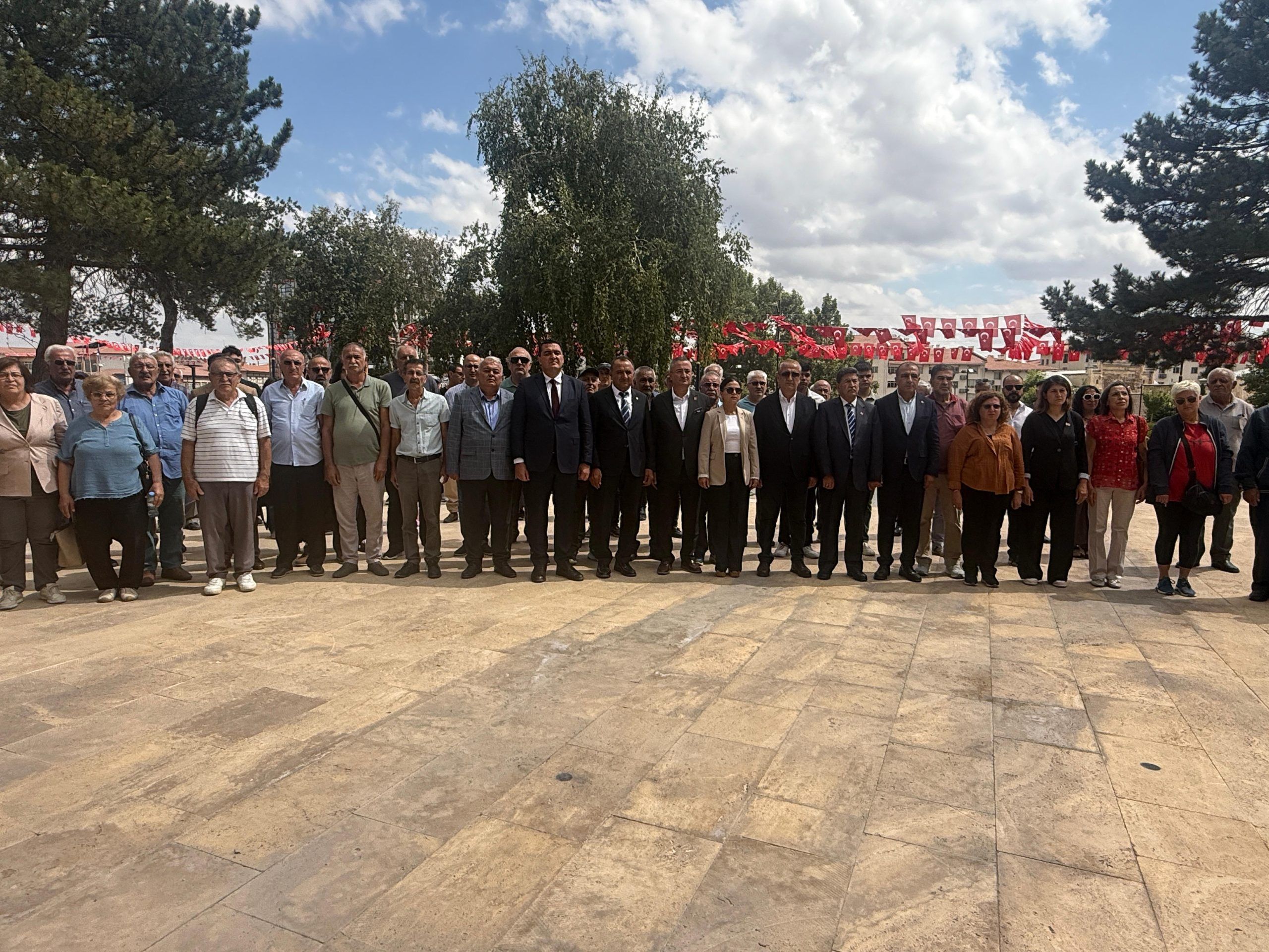 Açık havada, ağaçların önünde yan yana duran kalabalık bir grup ve arkalarında Türk bayrakları görülüyor.