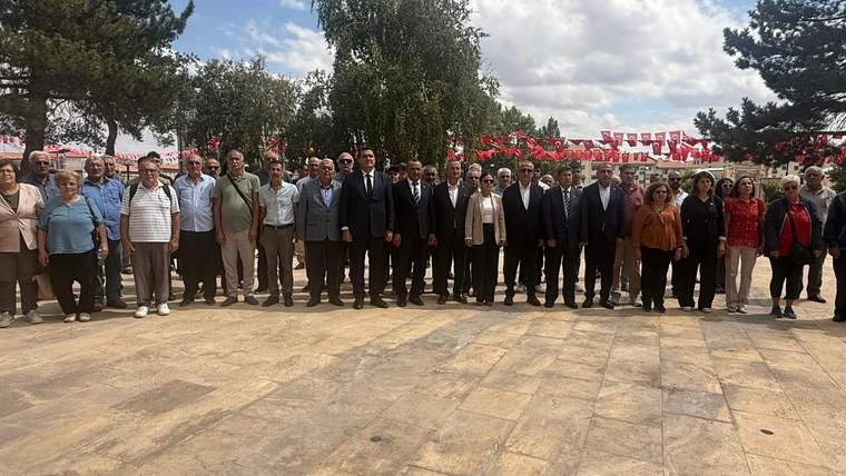 Açık havada, ağaçların önünde yan yana duran kalabalık bir grup ve arkalarında Türk bayrakları görülüyor.