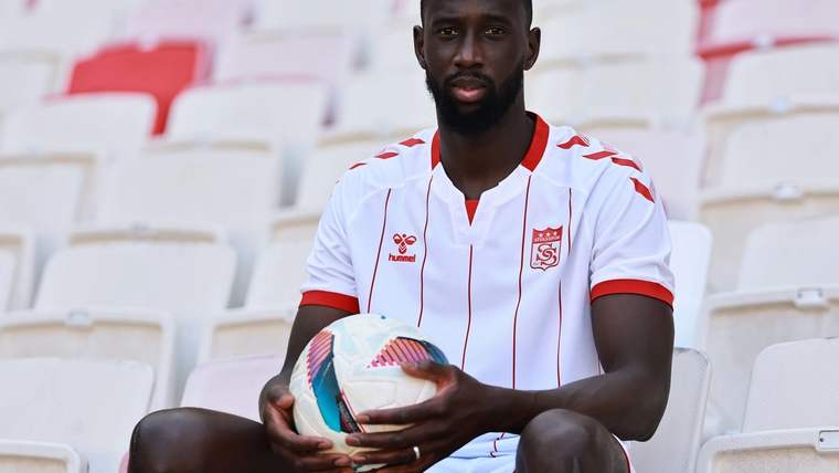 Kırmızı-beyaz forma giyen bir futbolcu, tribün koltuklarında elleriyle futbol topu tutuyor.