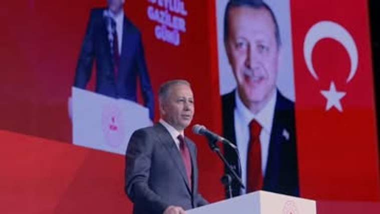 Kürsüde konuşan bir kişi, kırmızı fonda Türk bayrağı ve iki portre bulunan salonda görülüyor.