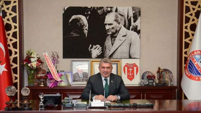 Bir masanın arkasında oturan bir kişi, masanın üzerinde çiçekler ve çeşitli nesneler var.