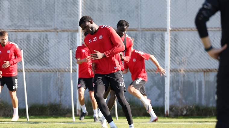 Kırmızı formalar giyen futbolcular, çim sahada antrenman sırasında koşu ve ısınma hareketleri yapıyor.
