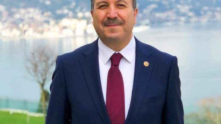 Siyah takım elbise giymiş bir kişi, arka planda su ve şehir manzarasıyla gülümsüyor.