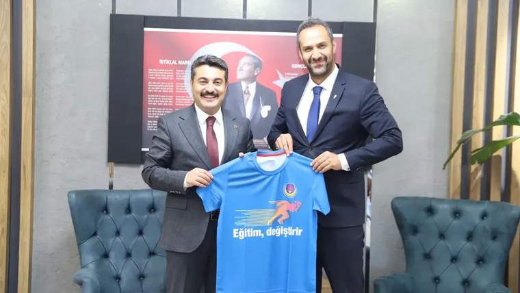 İki kişi, mavi bir tişört tutarak gülümsüyor. Arka planda Türk bayrağı ve bir poster var.