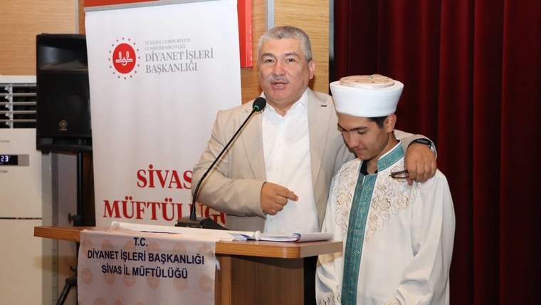 Mikrofonun önünde konuşan bir kişi, yanında geleneksel kıyafet giymiş bir çocuk duruyor.